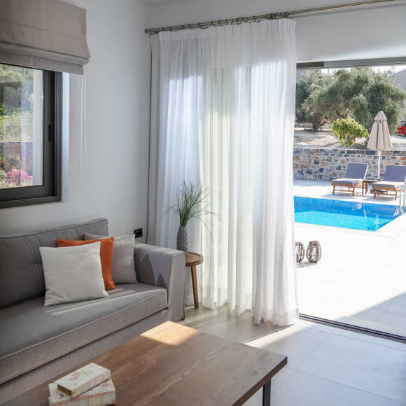Grand villa living room with grey sofa, coffee table and view to the pool through glass doors at Avdou Collection