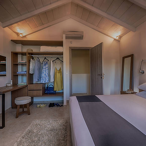 Junior villa bedroom with double bed, open wardrobe with hanging clothes, desk with stool and mirror at Avdou Collection